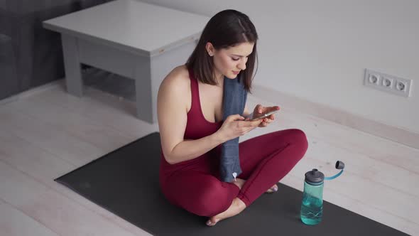Lady in Sports Wear Sitting on Yoga Mat with Crossed Legs and Using Smartphone After Trainings alt
