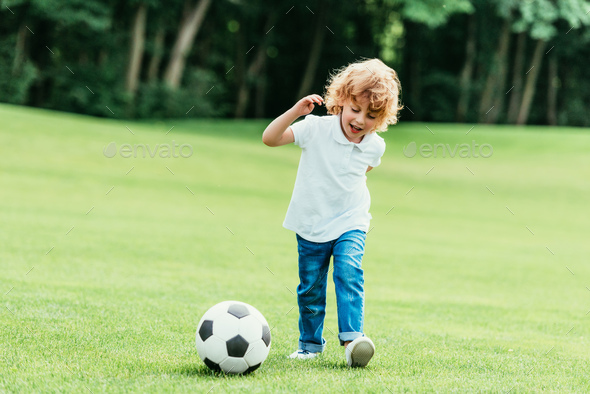 cheerful little boy playing with soccer ball on green lawn at park ...