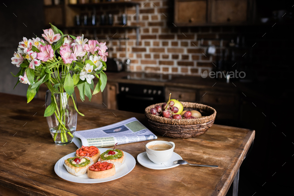 Still life of flowers, newspaper, breakfast with cakes, coffee and fruits on kitchen table
