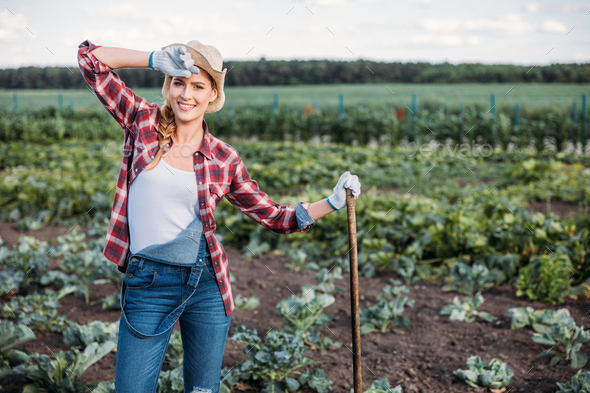 beautiful young female farmer in hat holding hoe and working on field