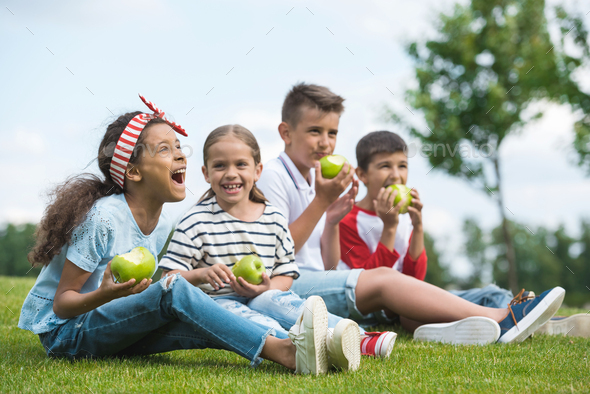 Happy multiethnic children eating green apples while sitting together on green grass
