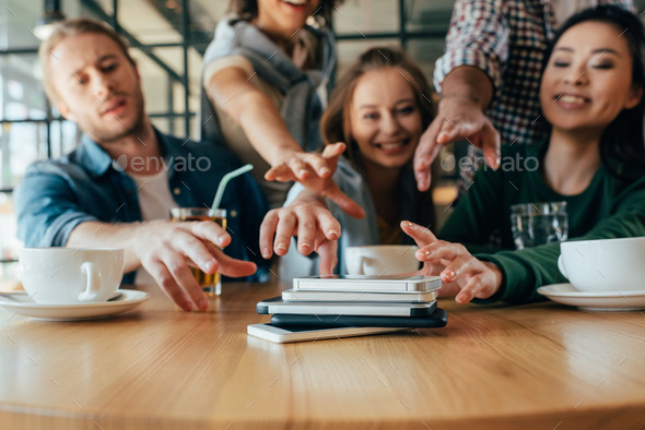 Young friends taking back their smartphones lying in pile on table in cafe
