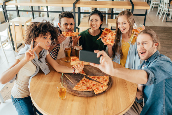 Young man taking selfie with multiethnic friends having pizza in cafe
