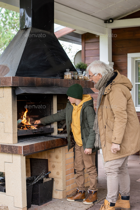 Cute boy in warm jacket and beanie standing by fireplace while putting firewoods