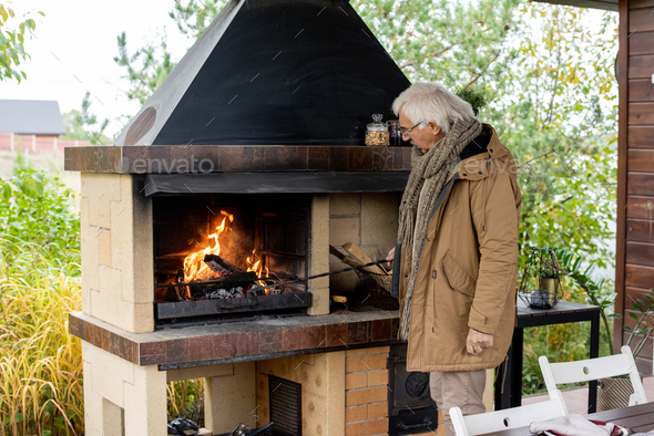 Contemporary senior man in warm casualwear standing by fireplace by house