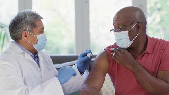 Male doctor wearing face mask vaccinating african american senior man at home alt