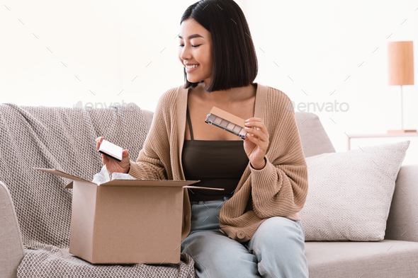 Beauty Box Concept. Portrait of smiling asian lady received delivery package, holding eyeshadow palette for makeup, unpacking cardboard parcel with cosmetics product, sitting on the couch at home