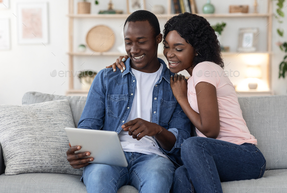 Joyful black family husband and wife using digital tablet, sitting on couch at living room, empty space. Smiling african american man and woman holding brand new pad, playing game online or websurfing