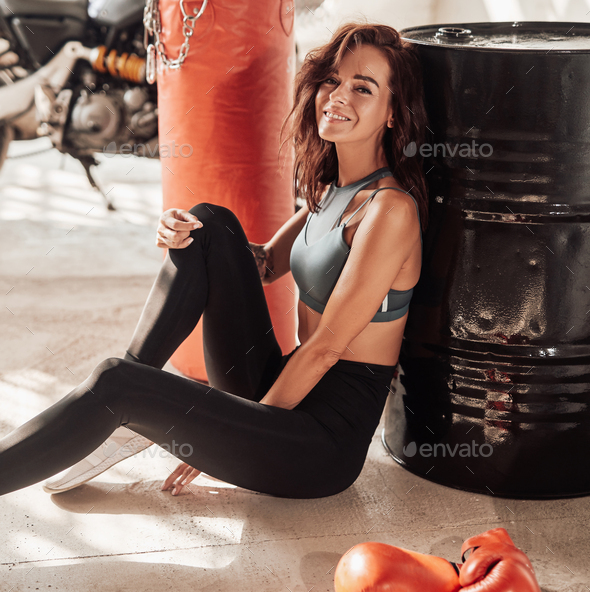 Slim female boxer sitting on floor leaning on black barrel with sport motorbike on background.