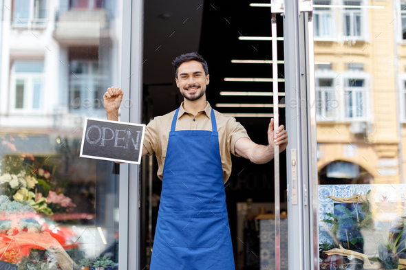 happy young florist in apron holding open sign and smiling at camera - Stock Photo - Images