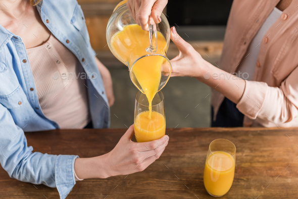 two women, senior and young drinking orange juice at table in kitchen. Woman pouring juice into