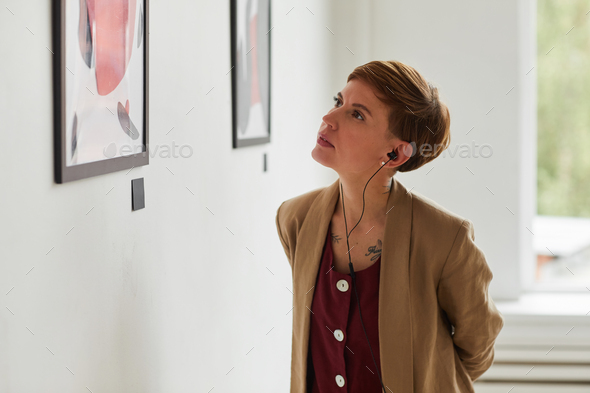 Waist up portrait of tattooed young woman looking at paintings and listening to audio guide at modern art gallery exhibition, copy space