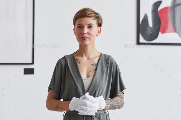 Waist up portrait of tattooed creative woman looking at camera while standing against white wall in modern art gallery exhibition, copy space
