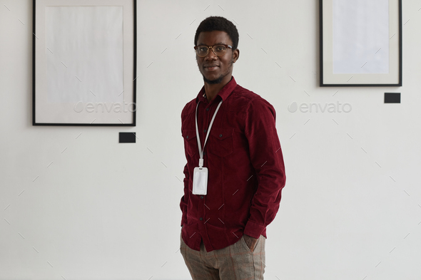 Portrait of young African-American man looking at camera with hands in pockets while working at art gallery or exhibition, copy space