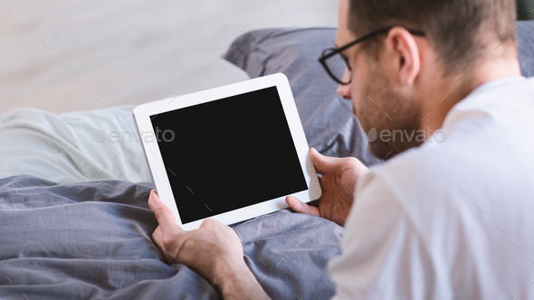 Man Using Digital Tablet With Empty Screen Browsing Internet Lying In Bed In Bedroom At Home. Computer Blank Display For Application And Website Advertisement Concept. Panorama, Mockup