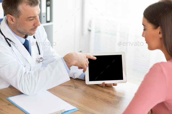 Medical Test And Examination Results. General practitioner showing patient digital tablet with black blank screen, discussing treatment, sitting at the table at office, pointing at monitor