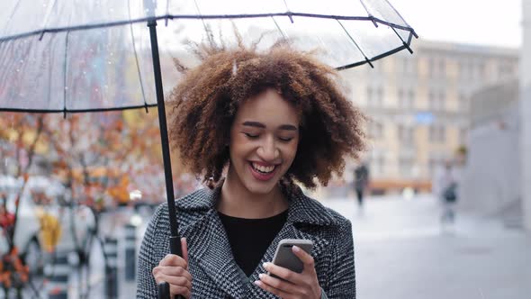 African American Girl Woman with Transparent Umbrella Stands in City in Rain Watching Funny Video on alt