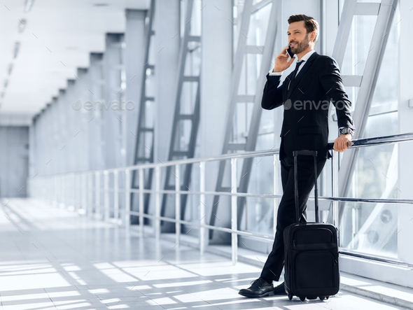Happy wealthy businessman standing in airport, talking on mobile phone