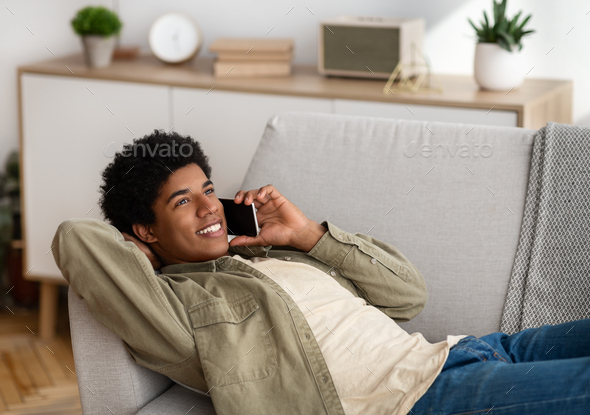 Funky African American teenager speaking on smartphone while lying on sofa at home, copy space. Handsome black guy having conversation with friend, family or teacher on mobile device indoors