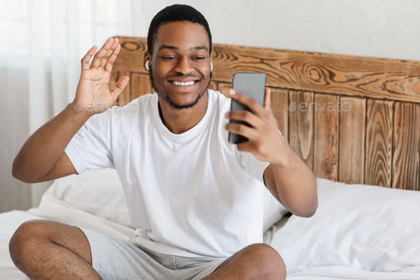 Cheerful African Guy Making Video Call On Cellphone, Waving Hello To Smartphone Sitting In Bed In Modern Bedroom At Home. Distant Communication, Mobile Online Video Conference App Concept