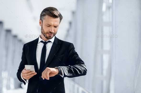 Just in time. Handsome bearded businessman checking time on his watch, using mobile phone while waiting for flight, walking by airport, copy space. Middle-aged entrepreneur using smartphone