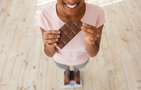 Diet failure, cheat meal and unhealthy nutrition. Above view of black woman holding chocolate on