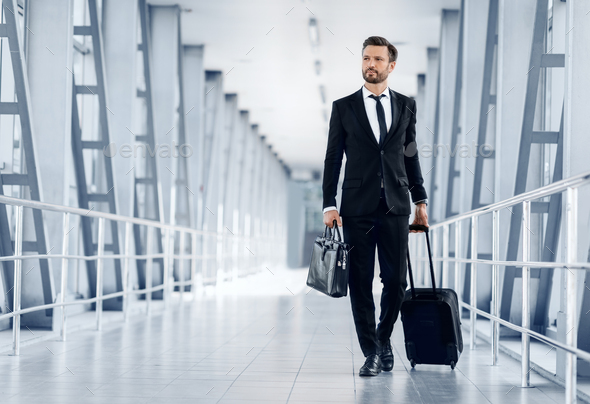 Middle-aged handsome businessman walking by airport terminal with luggage and case, looking at copy space. Rich entrepreneur in black suit going by modern airport, departuring for business trip