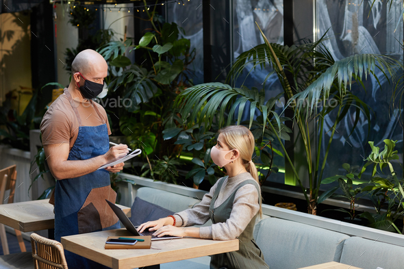Businesswoman in protective mask working at the table with laptop and making an order to the waiter in mask theey are in the restaurant