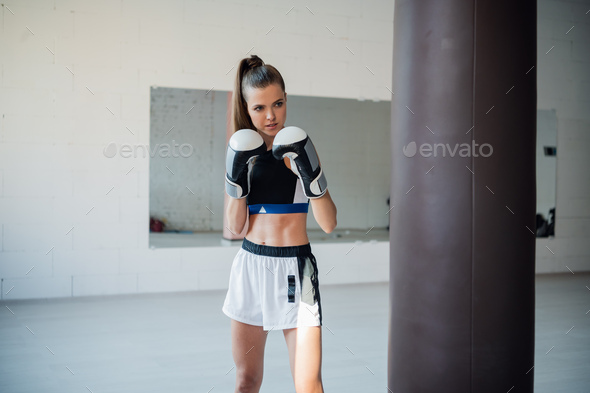 The girl is preparing for a boxing competition and trains punches on a punching bag in a spacious gym.