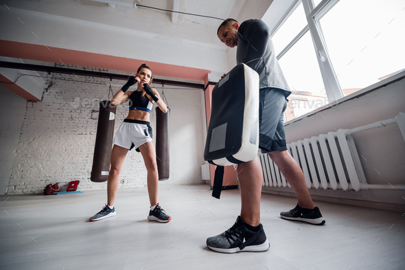 A young strong kickboxer girl kicks her sparring partner with a boxing paw in a personal training session.