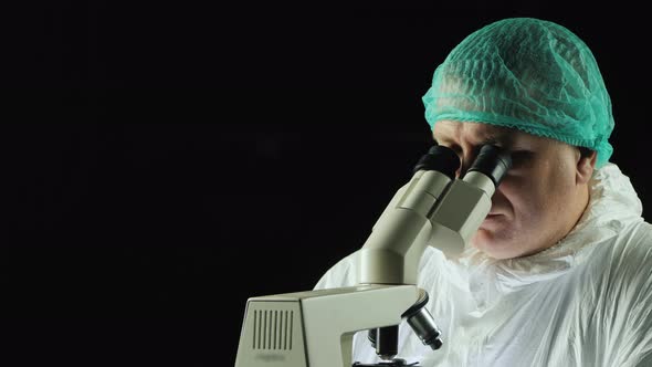 Male Lab Technician Looks Into Microscope Against Black Background alt