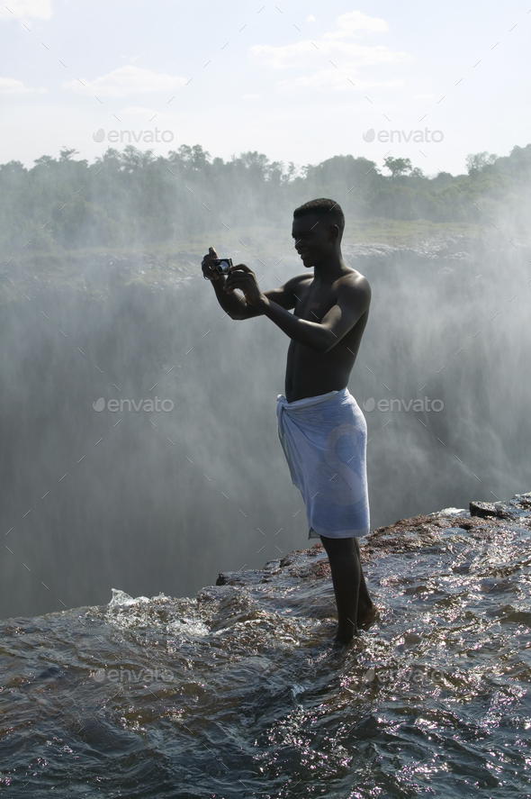 Man standing on ledge of Victoria Falls, taking picture, Zambia.