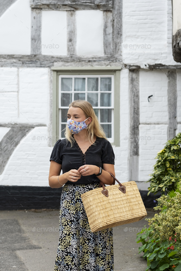 Young blond woman wearing face mask walking through village, carrying shopping bag. - Stock Photo - Images