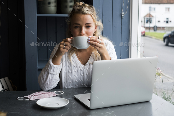 Young blond woman sitting alone at a cafe table with a laptop, and cup of coffee