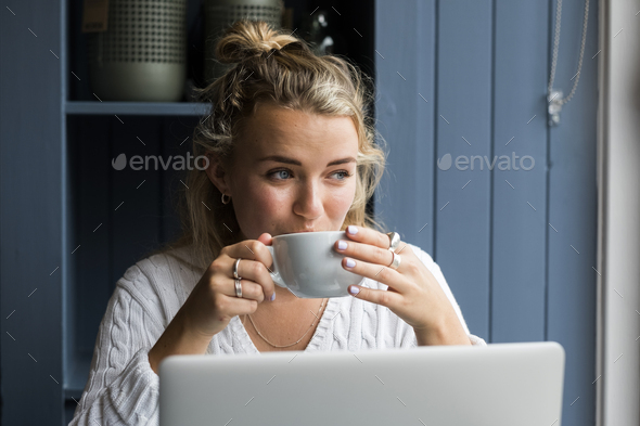 Young blond woman sitting alone at a cafe table with a laptop computer, working remotely.