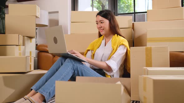 Asian woman with casual cloth sit among stack of boxes and use laptop with happiness