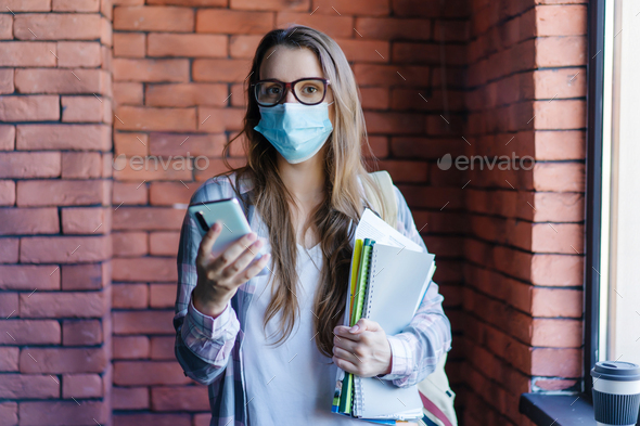 Female student wearing medical mask with textbooks and mobile phone in college.