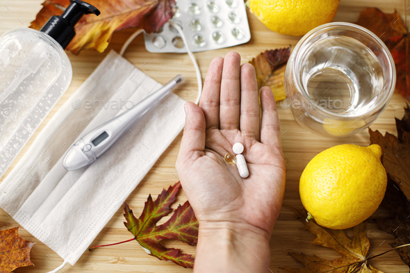 Hand with vitamin D, vitamin C and zinc pills on background of fall leaves, face mask, lemons