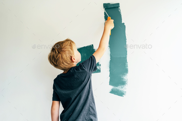 Caucasian blond boy paints a white wall with a roller in green. The child helps to make repairs.