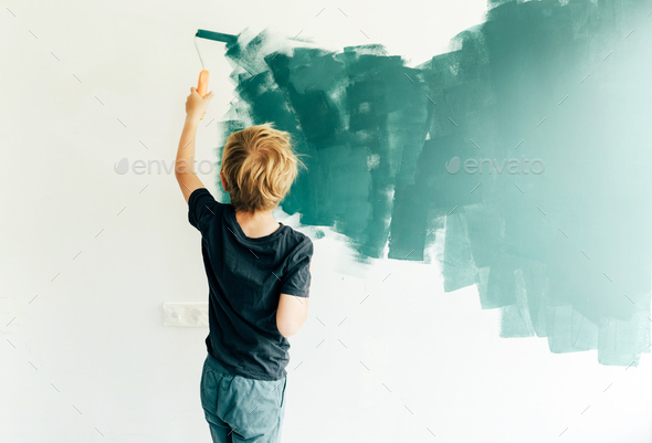 Caucasian boy paints a white wall with a roller in green. A schoolboy helps to make repairs.