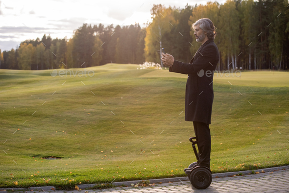 Side view of grey-haired businessman in elegant black coat making selfie by lawn - Stock Photo - Images