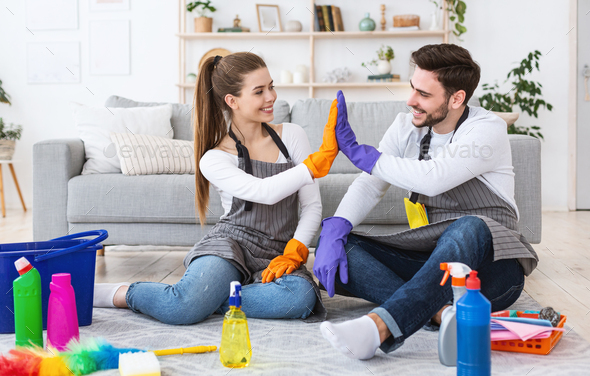 Smiling guy and woman in aprons and rubber gloves giving high five sitting on floor near cleaning