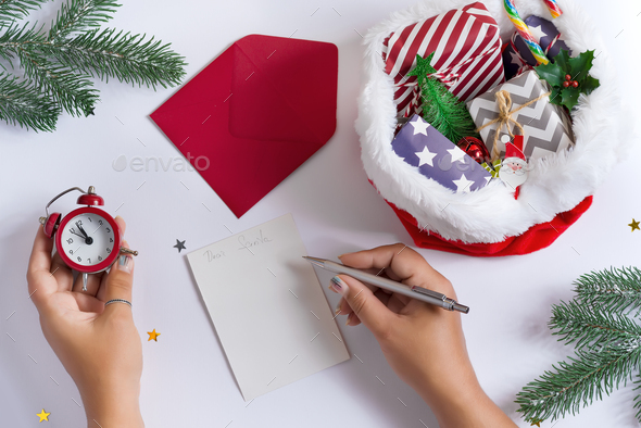 Christmas card with woman’s hands writing letter to Santa Claus, holding alarm clock and red hat filled different gifts on a light grey backgound, copy space. Top view. Congratulation postcard.