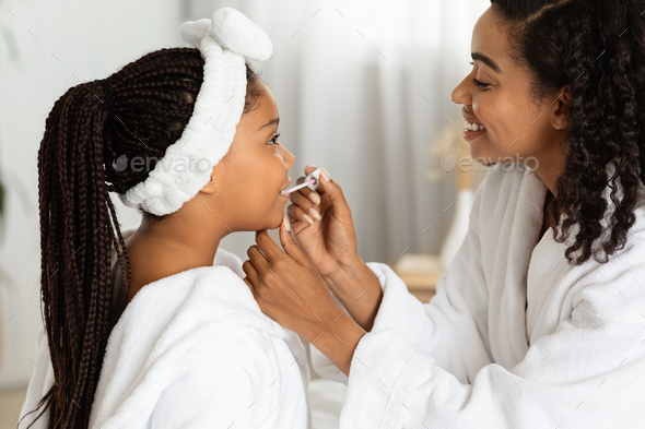 First Makeup. Caring Black Mom Applying Lip Gloss To Her Little Daughter