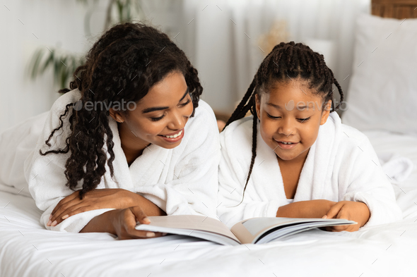 Little Black Girl And Mom Wearing Bathrobes, Lying On Bed With Magazine