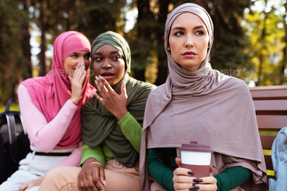 Muslim Ladies Whispering Behind Back Of Female Friend Sitting Outdoors