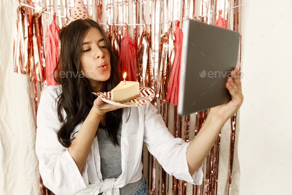 Happy woman making a wish and blowing candle on birthday cake and holding tablet with video call