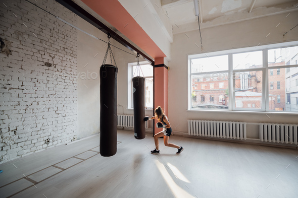 A girl in sports clothes is engaged in boxing and works out a punch with her hand on a punching bag.