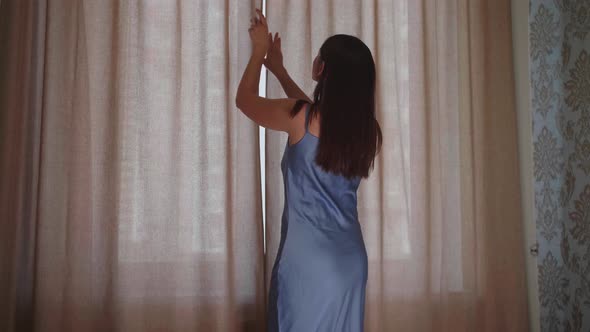 Young Woman Approaches the Window in the Bedroom Pushes Beige Curtains with Her Hands and Enjoys