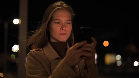 Woman Is Chatting in the Smartphone Against the Background of the Evening City alt